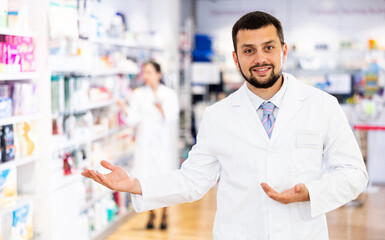 Portrait of positive man druggist standing in drugstore and making presenting gesture.