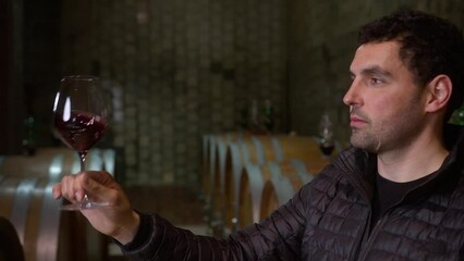 Caucasian man tasting red wine in a winery barrel cellar, holding a wine glass, swirling it, smelling, and sipping wine, delighted with wine taste and flavor looking at camera, close-up shot. - Powered by Adobe