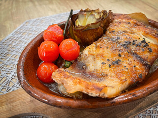 grilled corvine with roasted cherry tomatoes and grilled artichoke, in an oven clay pot on a wooden board, mediterranean cuisine