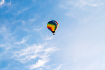 Rainbow colored hot air balloon flies in the blue sky with white cirrus clouds.