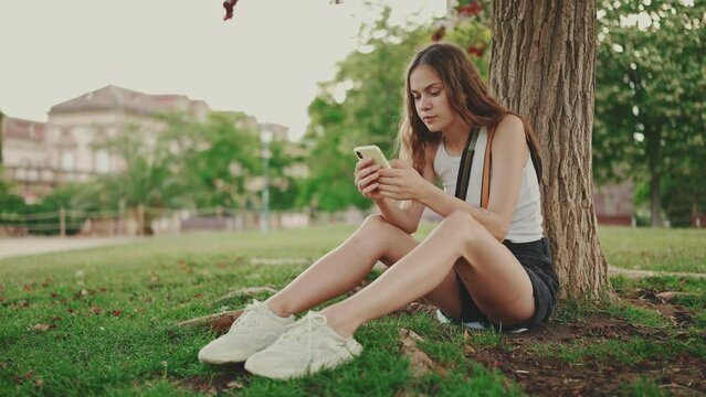 Beautiful girl with long wavy hair dressed in white top uses mobile phone while sitting on the green grass under tree in the park. Girl flip through the pictures on your phone with fingers