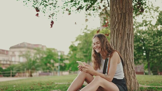 Beautiful girl with long wavy hair dressed in white top uses mobile phone while sitting on the green grass under tree in the park. Girl flip through the pictures on your phone with fingers