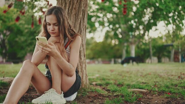 Beautiful girl with long wavy hair wearing in white top uses mobile phone while sitting on the green grass under tree in the park. Girl flip through the pictures on your phone with fingers