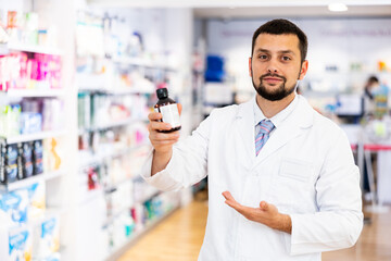 Portrait of confident young adult bearded man pharmacist offering product, working at pharmacy