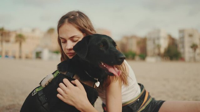 Beautiful girl with long wavy hair wearing white top is sitting on the sand with black dog on modern building background. Girl hugging and stroking her pet