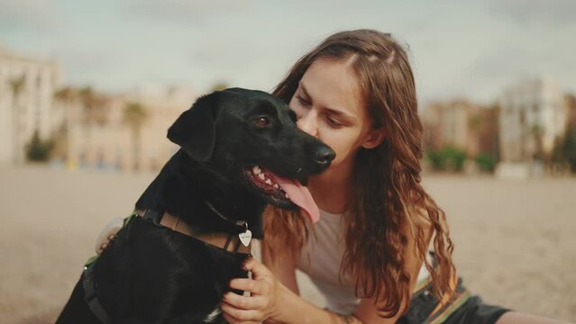 Clouse-up, beautiful girl with long wavy hair wearing white top is sitting on the sand with black dog on modern building background. Girl petting and kissing her pet