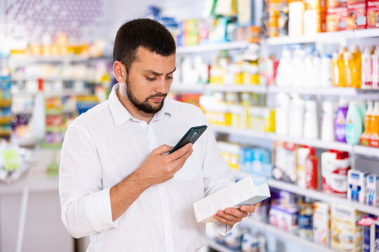 Portrait of focused bearded man choosing cosmetic product at pharmacy, using smartphone for scanning barcode