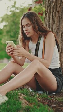 VERTICAL VIDEO: Beautiful girl uses mobile phone while sitting on the green grass under tree in the park. Girl flip through the pictures on your phone with fingers