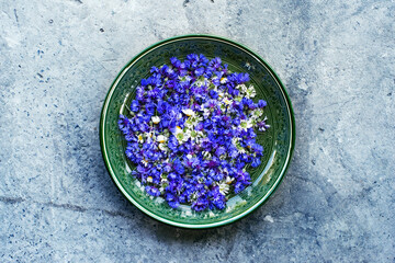 Obraz na płótnie Canvas Bright blue flowers of cornflowers and chamomile in a round ceramic green plate on gray grunge concrete surface, top view. Floral background with summer meadow grasses