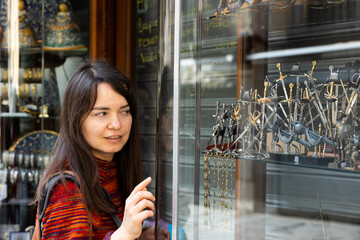 Young female tourist, standing near the window of a souvenir shop, carefully examines the goods,...