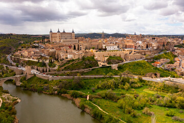 Naklejka premium Picturesque aerial view of Toledo cityscape on green hilly banks of Tagus river in early spring overlooking fortified Alcazar castle framed by four towers, Spain
