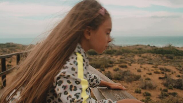 A little girl with a long hair climbing up on a deck on the beach on a cloudy day