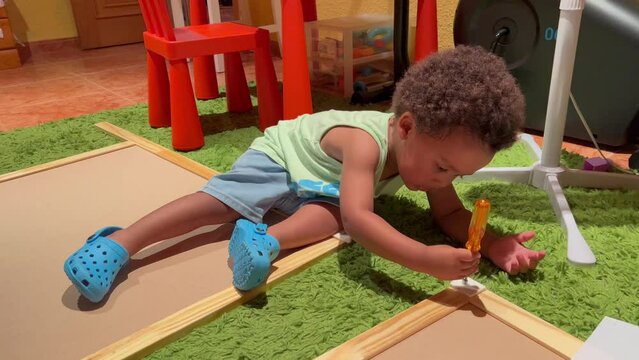 Adorable, smart and exotic two year old afro european child helping daddy to assemble a forniture trying to ude screws and a screwdriver. Wearing sleeveless green shirt and shorts, at home.