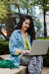 Caucasian woman smiling, sitting on a bench working on her computer.