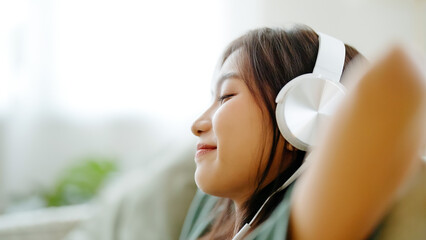 Young asian woman listening to music on couch in living room at home. Happy asia female using mobile smartphone, wearing headset and sitting on sofa