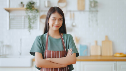 Happy young asian woman wearing apron and standing in kitchen room. Beautiful female smiling and...
