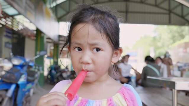 Happy Asian little girl eating popsicle at summertime, young woman child making a funny face as he is eating a frozen fruit popsicle outside, Kid having fun relaxing with ice cream