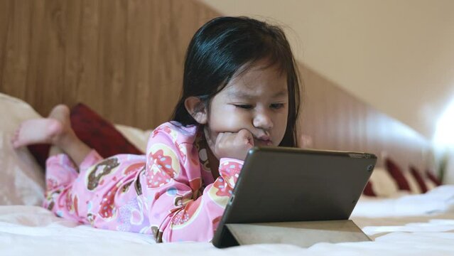 Kid watching videos and cartoons online in bedroom at home, Little Asian girl playing digital tablet on bed addicted game and cartoon, internet funny online concept