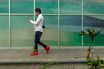 Young Latin American man walking, smiling, calm and relaxed attitude, with a cell phone in his hands, wearing a white shirt, blue jeans, camera hanging, in a multicolored background. 