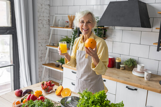 Attractive Senior Woman Drinking Freshly Squeezed Orange Juice, Looking Happy And Healthy. Concept Of Active Lifestyle Of Mature People, Cooking Organic Food. Mature Female At Home At Her Kitchen