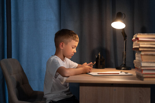 Distance learning online education. Caucasian kid boy studying at home with smartphone and doing school homework at night. Thinking child siting at table with cell phone. - Powered by Adobe