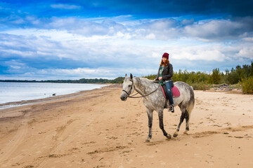 wearing jeans, jacket and warm hat female jokey rides astride grey horse along shore