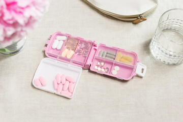 Top view various capsules and tablets in plastic container and glass of water on table. Pink pills box for daily intake of vitamins and medicines on cloth. Lifestyle for beauty and health