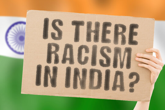 The question " Is there racism in India? " is on a banner in men's hands with a blurred Indian flag in the background. Interracial. Issues. Justice. Nation. Politics. Peace. Patriotism. Rebellion