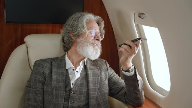 Senior male millionaire talking on the speakerphone or recording a voice message on his smartphone while flying on a private jet. Rich man talking on mobile phone while flying at first class airplane