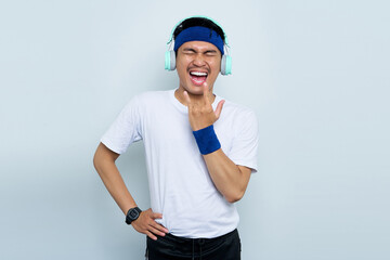 Crazy young Asian man sporty fitness trainer instructor in blue headband and white t-shirt. Listen music with headphones,  Depicting heavy metal rock sign isolated over white background