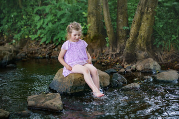 Cute little toddler girl having fun by a river on warm and sunny summer day. Happy excited preschool child splashing with water in forest stream creek.