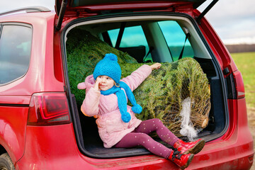 Adorable little preschool girl with Christmas tree inside of family car. Happy healthy child in winter fashion clothes choosing and buying big Xmas tree for traditional celebration.