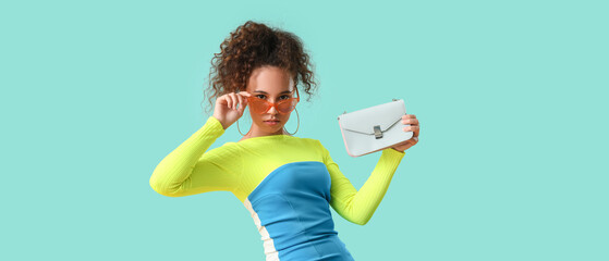 Beautiful young African-American woman with stylish bag on turquoise background