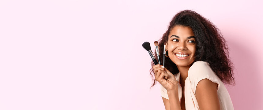Beautiful young African-American woman with makeup brushes on pink background with space for text