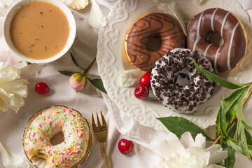 Top view of breakfast in bed with various delicious glazed donuts on the plate, cup of coffee and sweet cherry berries.