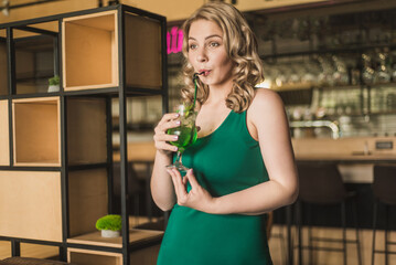 Fototapeta na wymiar Young plus size woman in a green dress with a cocktail on the background of the bar