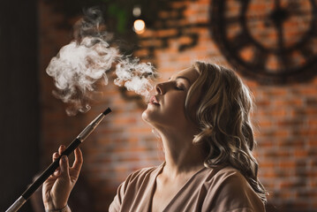 A young plus size  woman in a bar smokes a hookah and lets out smoke. Relax in the evening in the...