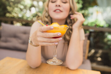 Young chubby pretty blonde woman with curls is drinking a summer cocktail in orange color on the terrace in a cafe. Plus size girl. Selective focus and glass close up
