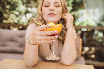 Young chubby pretty blonde woman with curls is drinking a summer cocktail in orange color on the terrace in a cafe. Plus size girl. Selective focus and glass close up