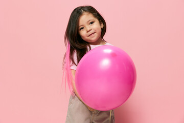 a beautiful little girl of preschool age stands on a pink background and a pink T-shirt and plays with a pink ball, hiding and peeking out from behind it, spreading her arms in different directions