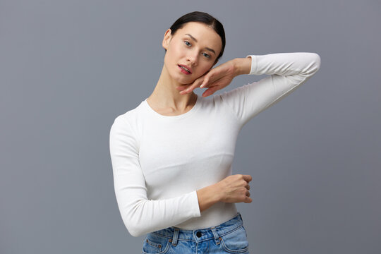a cute woman with a ponytail on her head stands on a dark background in a white tight T-shirt, put her hand on her head, and put the other on her stomach