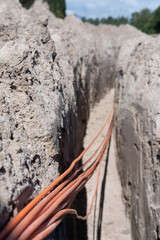 Bundle of orange fiber optic cables lies in a trench dug in the ground in a street. Focus on the cables at the bottom left, blurred background of the thrown sand