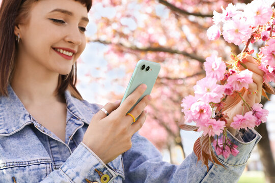 Beautiful young woman taking picture of blossoming sakura tree branch in park, focus on hand