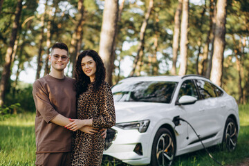 Young couple man and woman traveling together by new car having stop at charging station boyfriend holding plug playing gun looking at girlfriend holding cup drinking hot coffee laughing cheerful