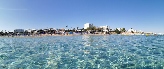 Protaras. Famagusta area. Cyprus. Panorama of Fig Tree Bay beach, people sunbathing and swimming, hotel buildings behind the beach against the sky with clouds. View from the sea.