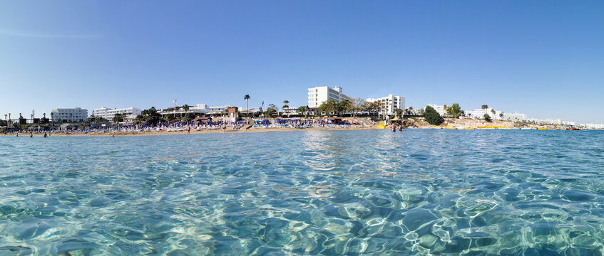 Protaras. Famagusta area. Cyprus. Panorama of Fig Tree Bay beach, people sunbathing and swimming, hotel buildings behind the beach against the sky with clouds. View from the sea.