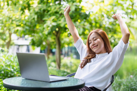 Portrait image of a woman with closed eyes, stretching arms and relaxing while working on laptop computer in the park