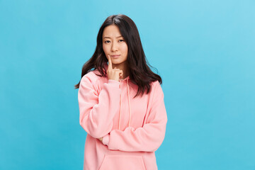 Confused pensive cute Asian student young lady in pink hoodie sweatshirt looks aside posing isolated on over blue studio background. The best offer for ad. People Emotions for Everyday concept