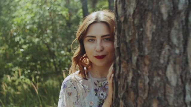 Portrait of beautiful young caucasian woman with red lips posing in the forest near tree bark, handled shot, view through tree branches.