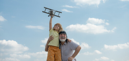 Grandson child and grandfather with toy plane over blue sky and clouds background. Two men generation grandfather and grandson playing outdoors. Generational family.